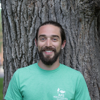 nick in a light green tee shirt smiling with a tree bark in the background