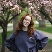 tatum in a blue sweatshirt smiling with hands on hips in front of a pink flowering tree